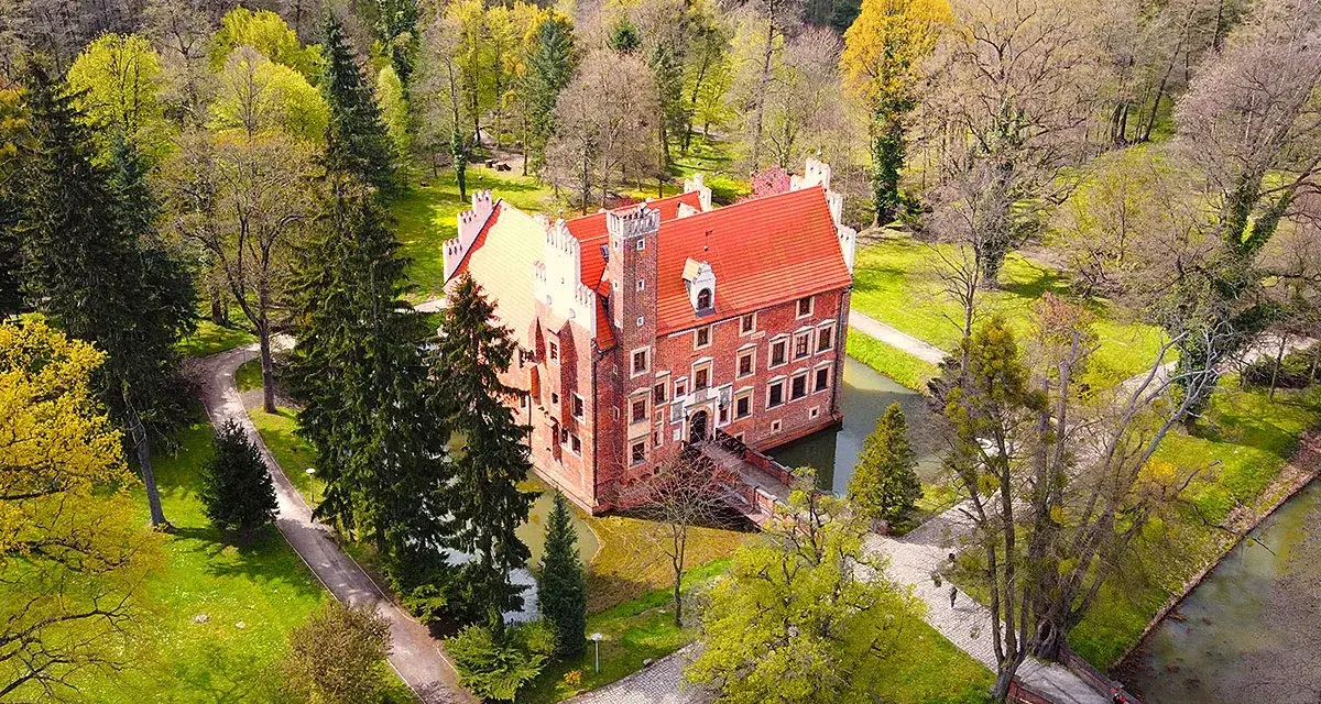 Zamek na wodzie w Wojnowicach otoczony zielenią i drzewami. Czerwona bryła zamku z charakterystycznym dachem wznosi się nad fosą.