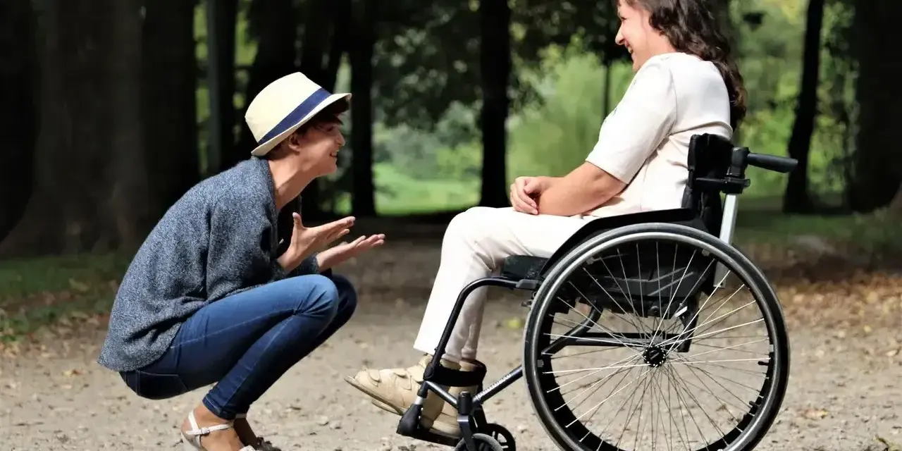 Opiekun osoby niepełnosprawnej pochyla się, by porozmawiać z kobietą na wózku inwalidzkim w parku.
