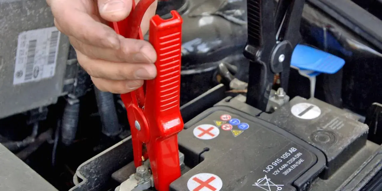 Hand klemmt rote Überbrückungsklemme an die Autobatterie, um Plus und Minus zu verbinden.
