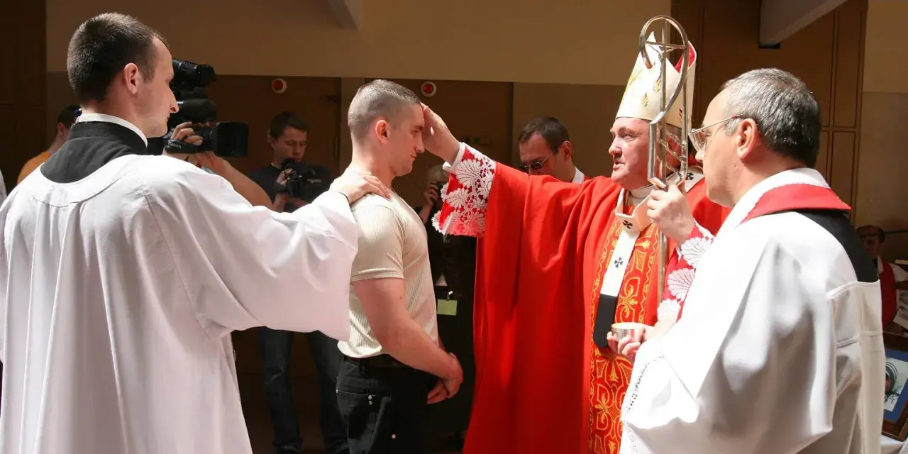 Biskup udziela sakramentu bierzmowania młodemu mężczyźnie. W tle widać osoby z kamerami.