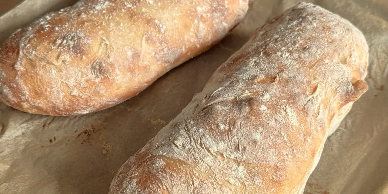 Dwa świeżo upieczone bochenki ciabatty, posypane mąką, leżą na papierze do pieczenia. Idealne do wypróbowania tego ciabatta przepis!