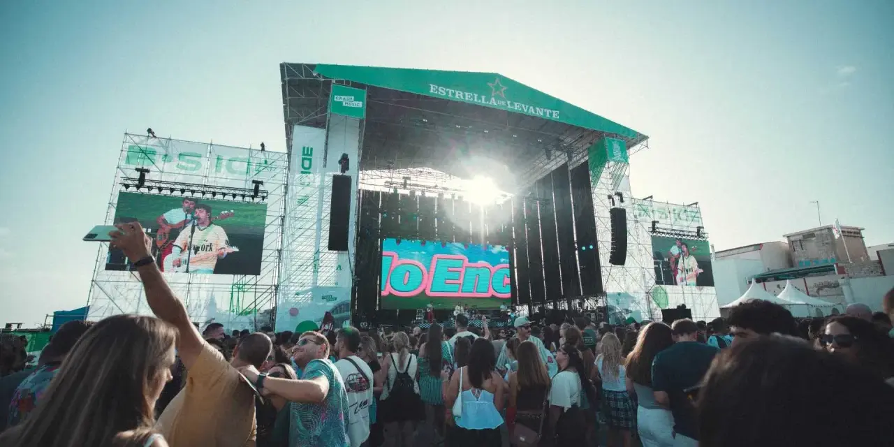 Multitud en concierto, con pantallas gigantes mostrando a artistas. El sol brilla sobre el escenario "Estrella de Levante" del festival B-Side.