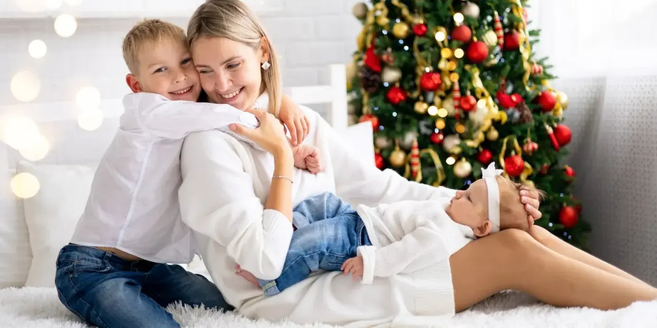 Rodzina cieszy się magicznymi świętami Bożego Narodzenia w hotelu. Syn mocno przytula mamę, a malutka córeczka śpi na jej kolanach.