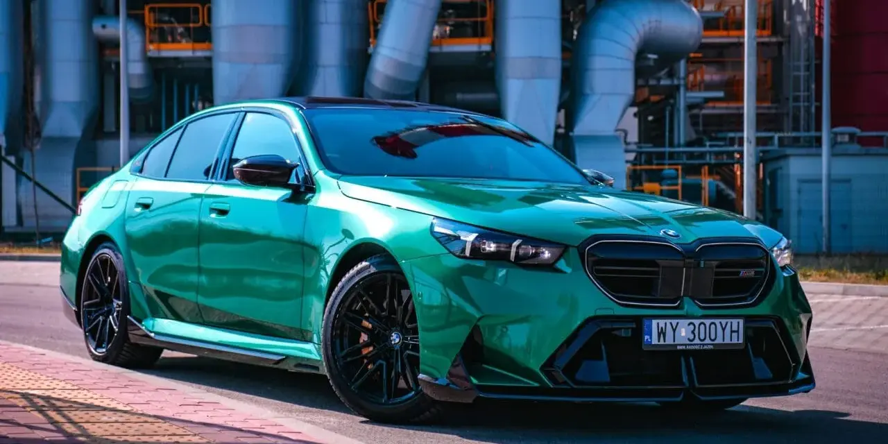 Błyszczące, zielone BMW M5 G90 z czarnymi felgami stoi na tle przemysłowych rur.