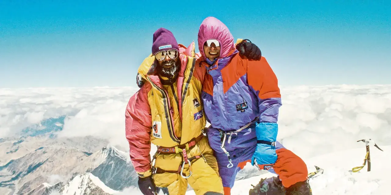 Zwei Bergsteiger auf dem Gipfel, die sich umarmen. Sie tragen bunte Daunenanzüge und Mützen. Die Frage "wie hoch ist der Mount Everest" wird hier beantwortet: 8848 Meter.
