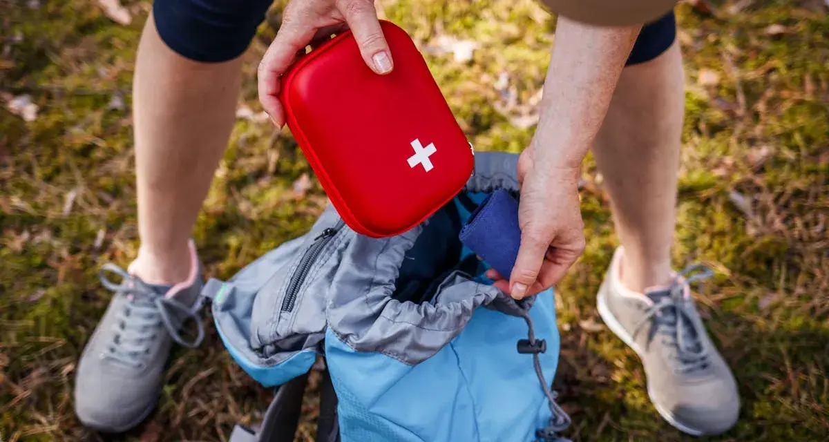Une personne prépare son sac à dos pour une randonnée, rangeant une trousse à pharmacie enfant rouge avec une croix blanche.
