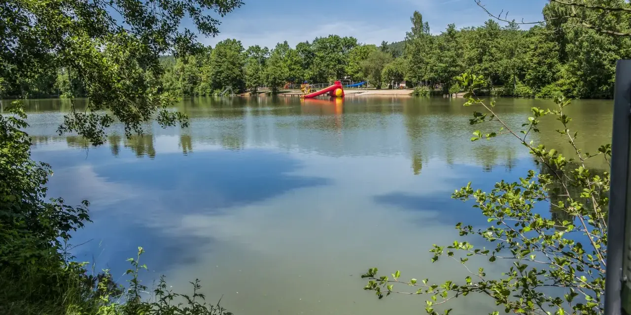 Idyllischer sulzfeld badesee mit Spielplatz und Rutsche, umgeben von grünen Bäumen.