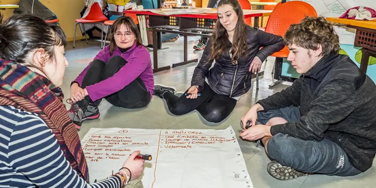 Quatre jeunes discutent autour d'un papier, participant à un mouvement d'éducation populaire. Ils brainstorment des idées pour améliorer une activité.