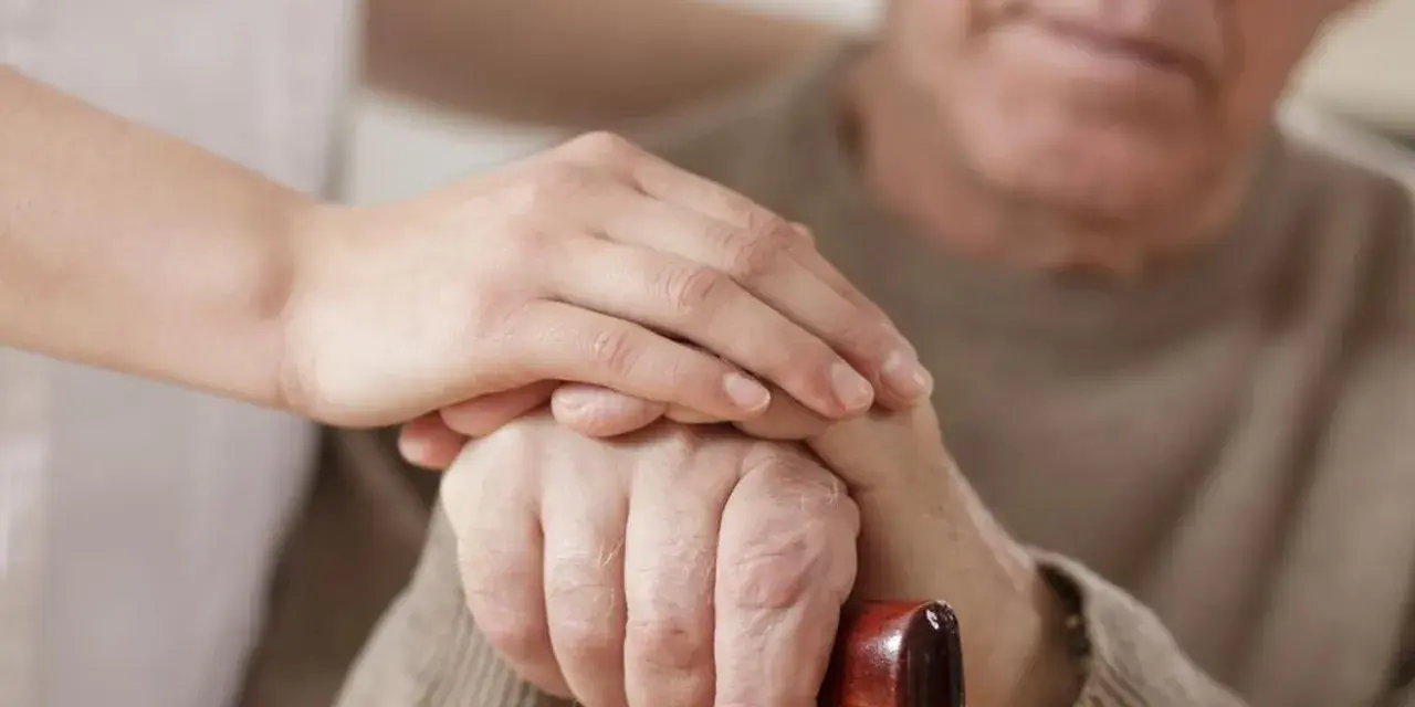 Dłoń opiekunki na dłoni starszego mężczyzny, który trzyma laskę. Wsparcie i troska, jak przy leczeniu np. chorobą Parkinsona, gdzie **Madopar** może pomóc.