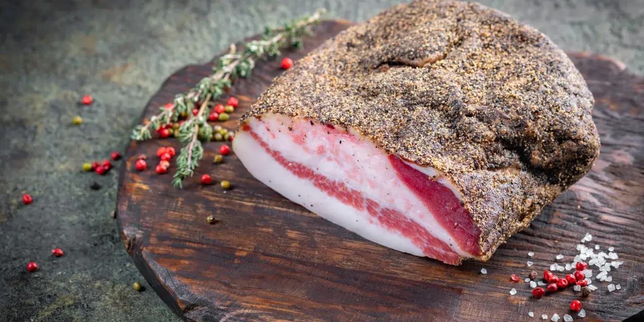 Guanciale co to? Włoski, peklowany policzek wieprzowy, pokryty pieprzem, leży na drewnianej desce z ziołami i pieprzem.