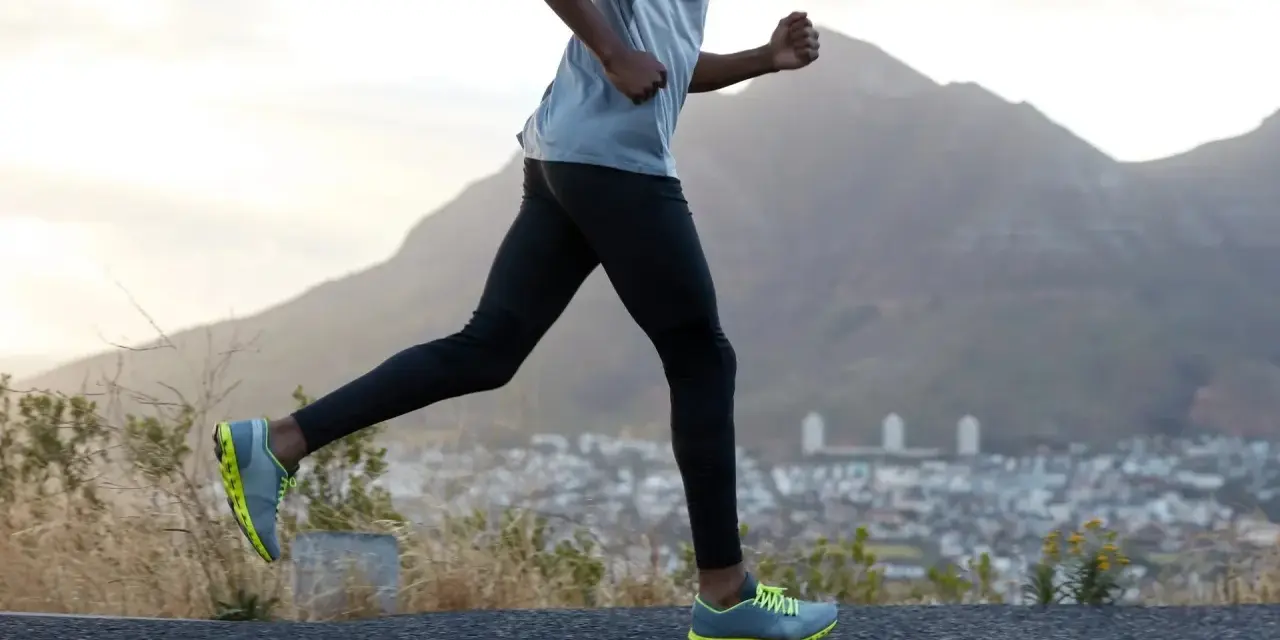 Un homme en tenue de sport fait du cardio training en courant sur une route de montagne, avec une ville en contrebas et des montagnes en arrière-plan.
