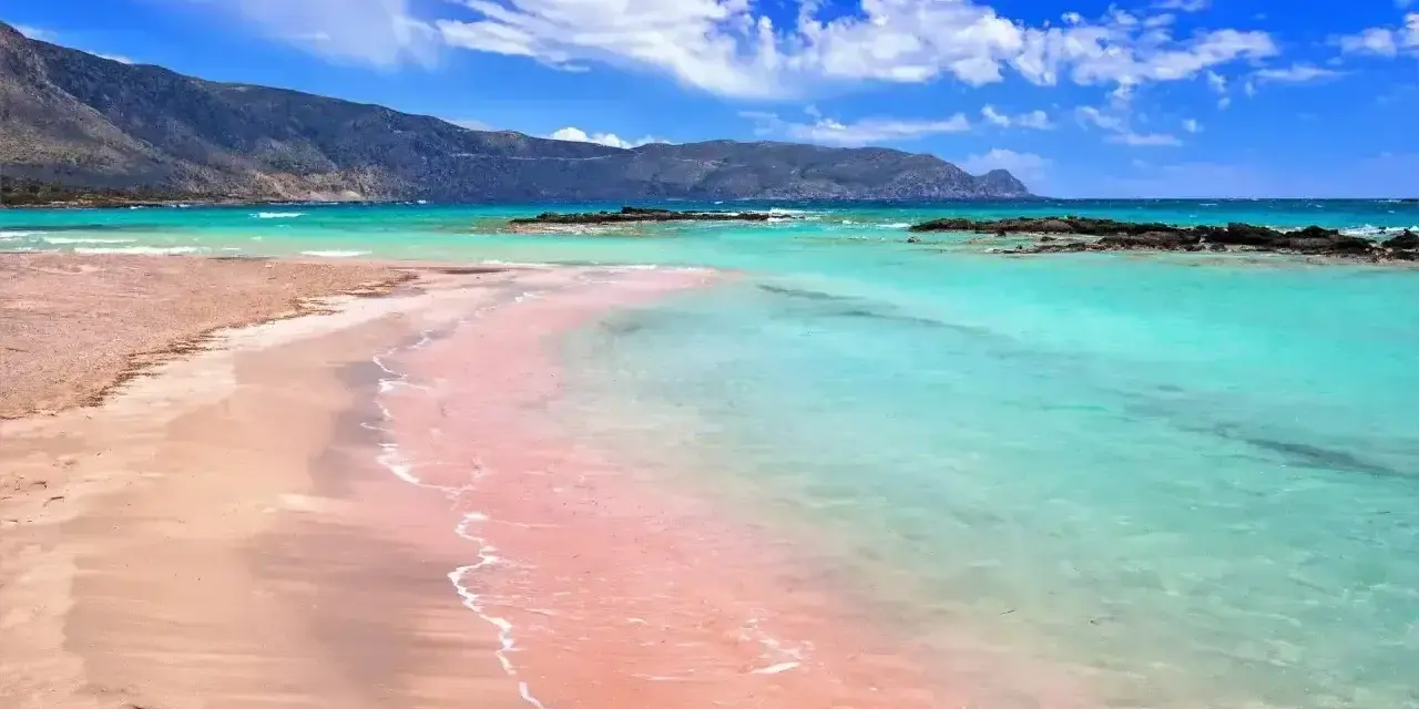 Różowy piasek plaży Elafonisi w Grecji, turkusowa woda i góry w tle.