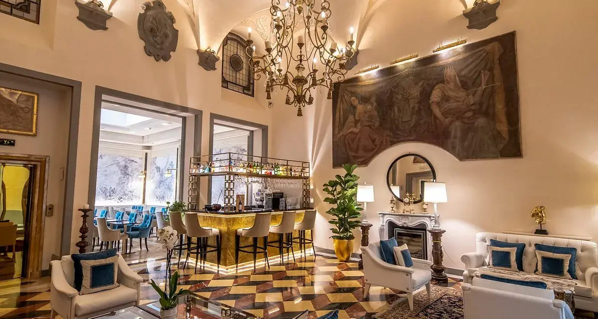Elegant hotel lounge with a bar, fireplace, ornate chandelier, and artwork.