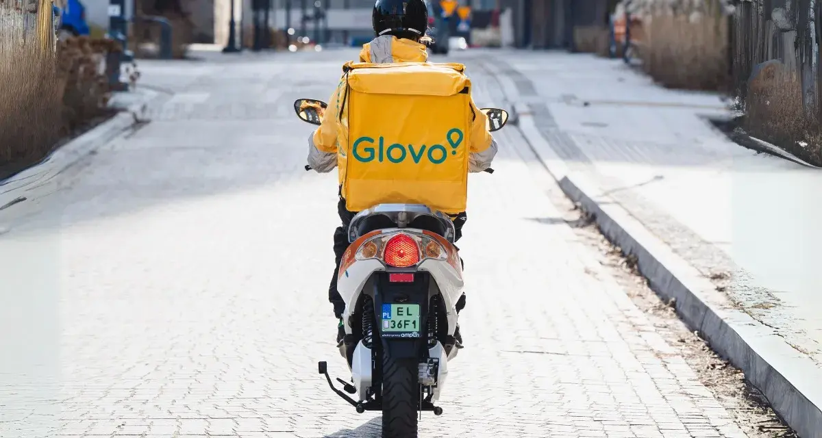 Kurier na skuterze z żółtym plecakiem Glovo jedzie ulicą.