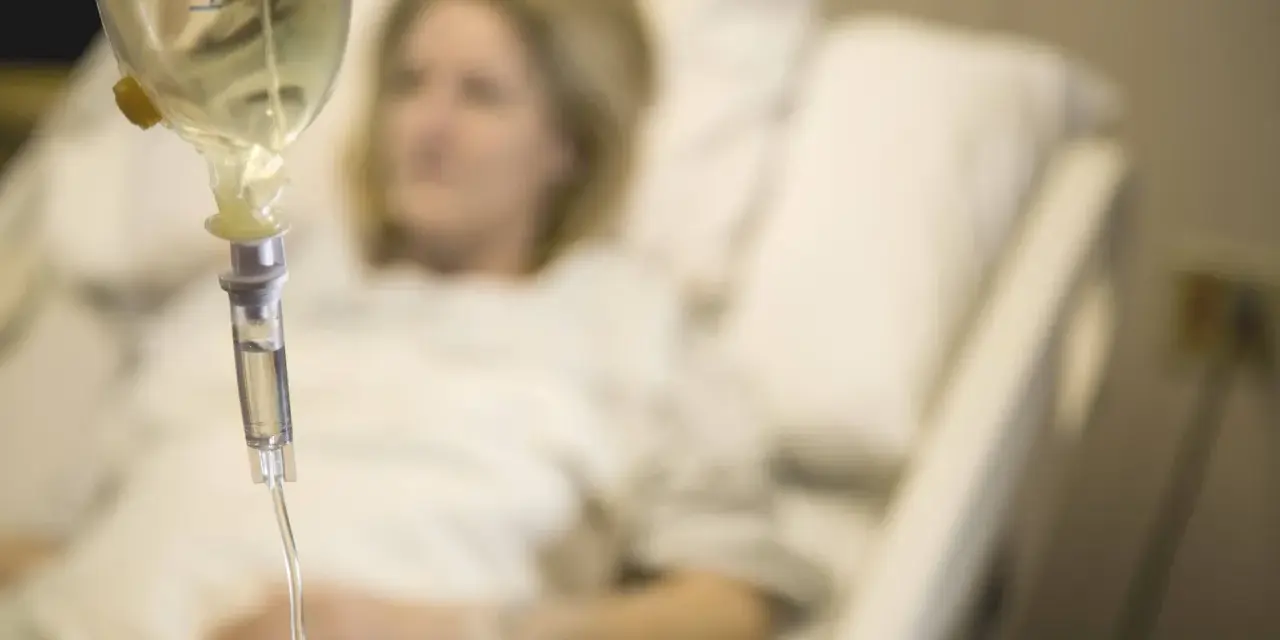 Infusion vorbereiten: Eine Frau liegt im Krankenhausbett, während eine Infusionstropfflasche und ein Tropfschlauch im Vordergrund zu sehen sind.