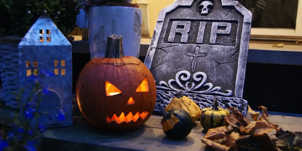 En läskig pumpa lyser upp med sitt ansikte, bredvid en gravsten och höstlöven. Perfekt för en kuslig bål halloween-kväll.