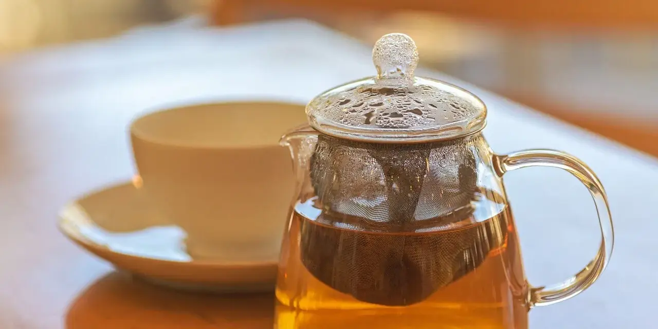 Théière en verre remplie de tisane dorée, avec une tasse en arrière-plan. La question est : combien de temps peut on garder une tisane infusée ?