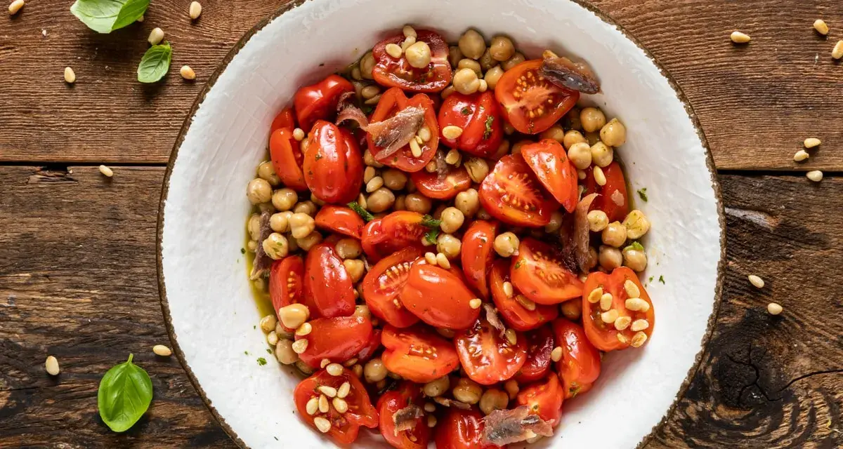 Insalata di ceci con pomodorini, acciughe e pinoli, guarnita con basilico fresco su un tavolo di legno rustico.