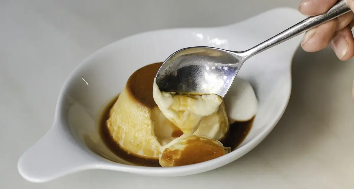 Una cuchara saca un trozo cremoso de flan de cuajada, bañado en caramelo líquido.