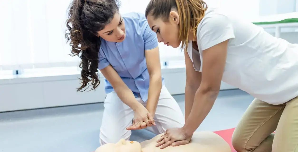 Zwei Frauen üben kardiopulmonale Reanimation an einer Übungspuppe. Eine Frau gibt Anweisungen, während die andere die Herzdruckmassage durchführt.