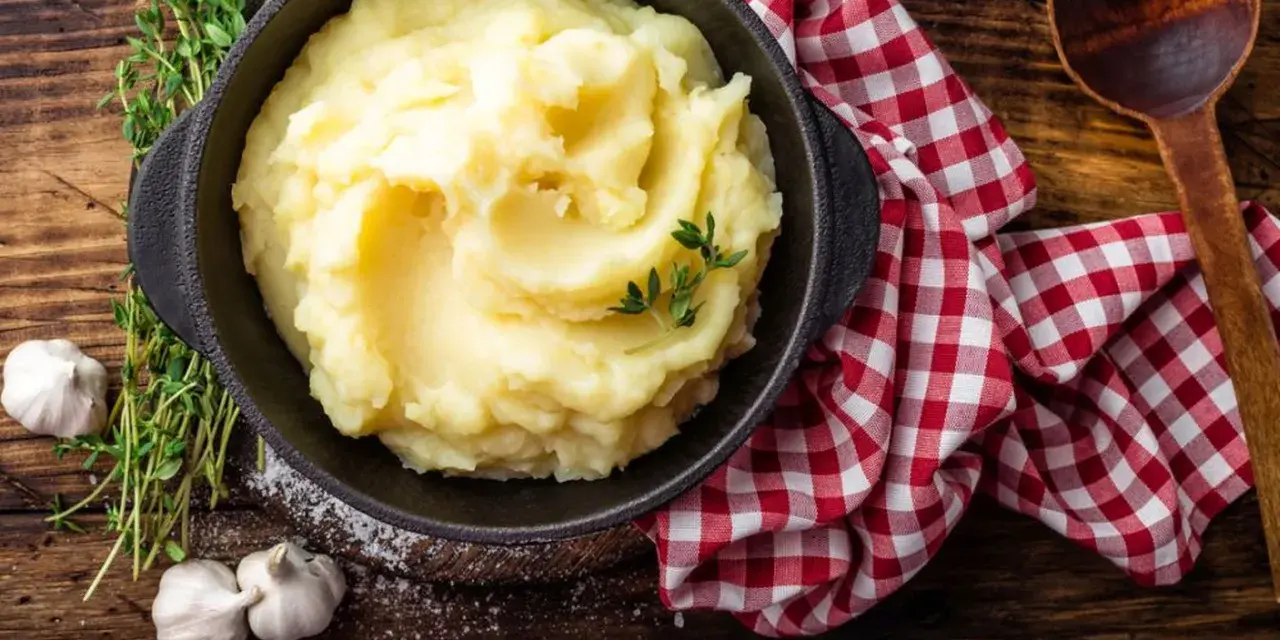 Una ciotola di cremoso purè di patate, guarnito con timo fresco, accanto ad aglio e un tovagliolo a quadri rossi.