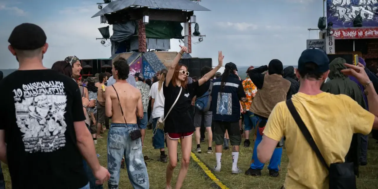 Une foule danse sous la pluie à un festival, une femme au centre lève les bras. On aperçoit une scène et des bannières, peut-être un dj français anime l'événement.