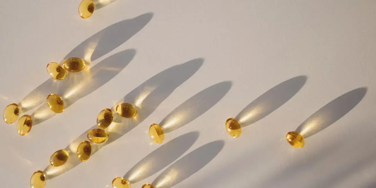 Gula kapslar med d-vitamin och magnesium sprids ut på en ljus yta, med långa skuggor som skapas av solljuset.