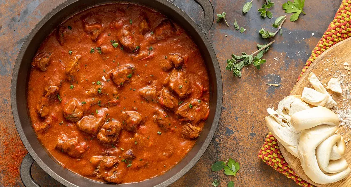 Un saporito gulash di manzo in una padella, accompagnato da pane fresco e erbe aromatiche.