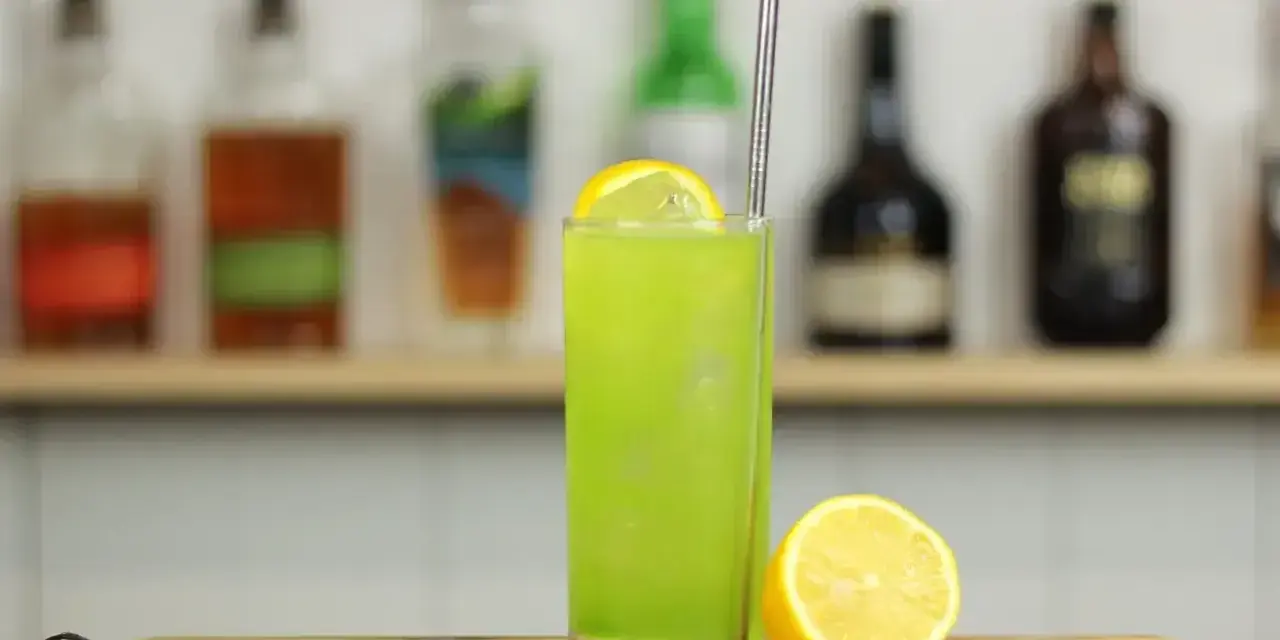 Zielony, orzeźwiający drink Tokyo Iced Tea z plasterkiem cytryny i metalową słomką, na tle półki z butelkami alkoholu.