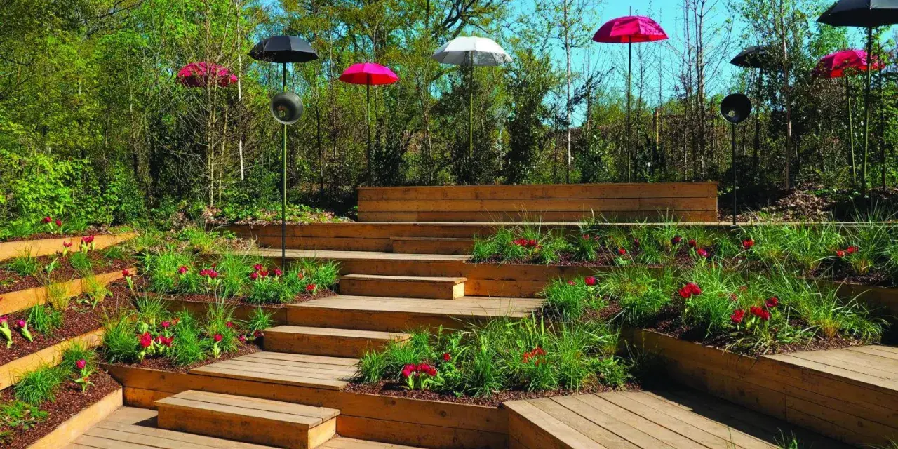 Un jardin en pente moderne avec des terrasses en bois, des parterres de fleurs rouges et des parasols colorés.