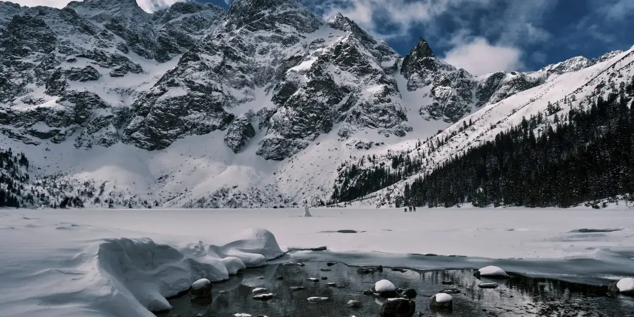 Zamarznięte Morskie Oko, otoczone ośnieżonymi szczytami Tatr. W oddali widać ludzi spacerujących po lodzie, a na pierwszym planie płynie strumyk.