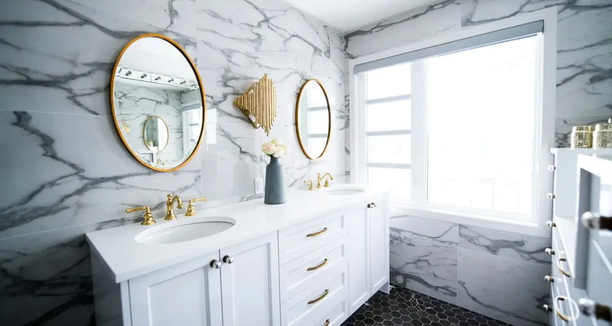 A modern bathroom vanity installation featuring white cabinets, marble walls, gold accents, and dual sinks.