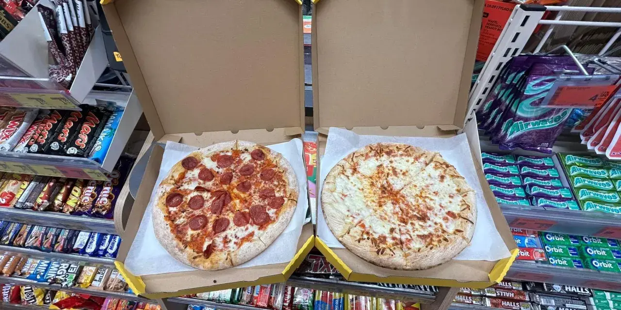 Dwie pizze w pudełkach, jedna z salami, druga serowa. Obok półki ze słodyczami i napojami w sklepie Żabka.