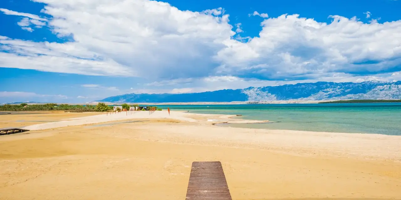 Drewniany pomost prowadzi na piaszczystą plażę Ninskiej laguny. W tle turkusowe morze i góry.