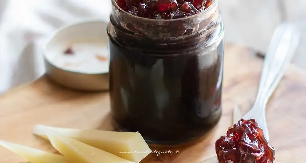 Un vasetto di marmellata di cipolle scura, con un cucchiaino che ne raccoglie una porzione, accanto a fette di formaggio stagionato.