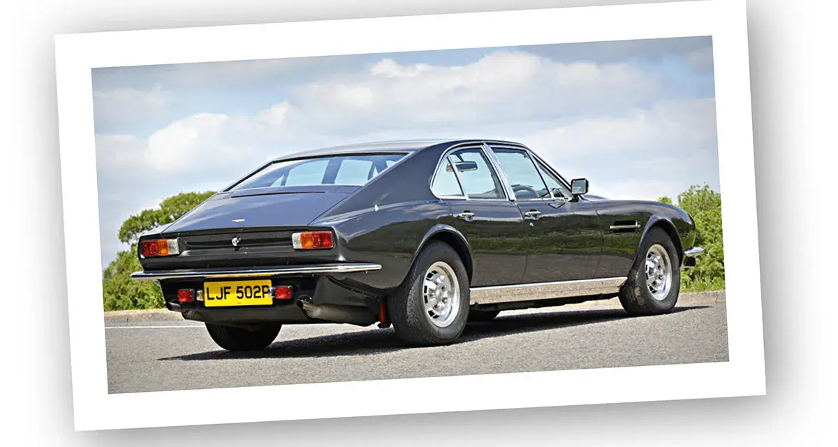 Klasyczny Aston Martin Lagonda w ciemnym kolorze, widziany z tyłu, na tle błękitnego nieba z chmurami.