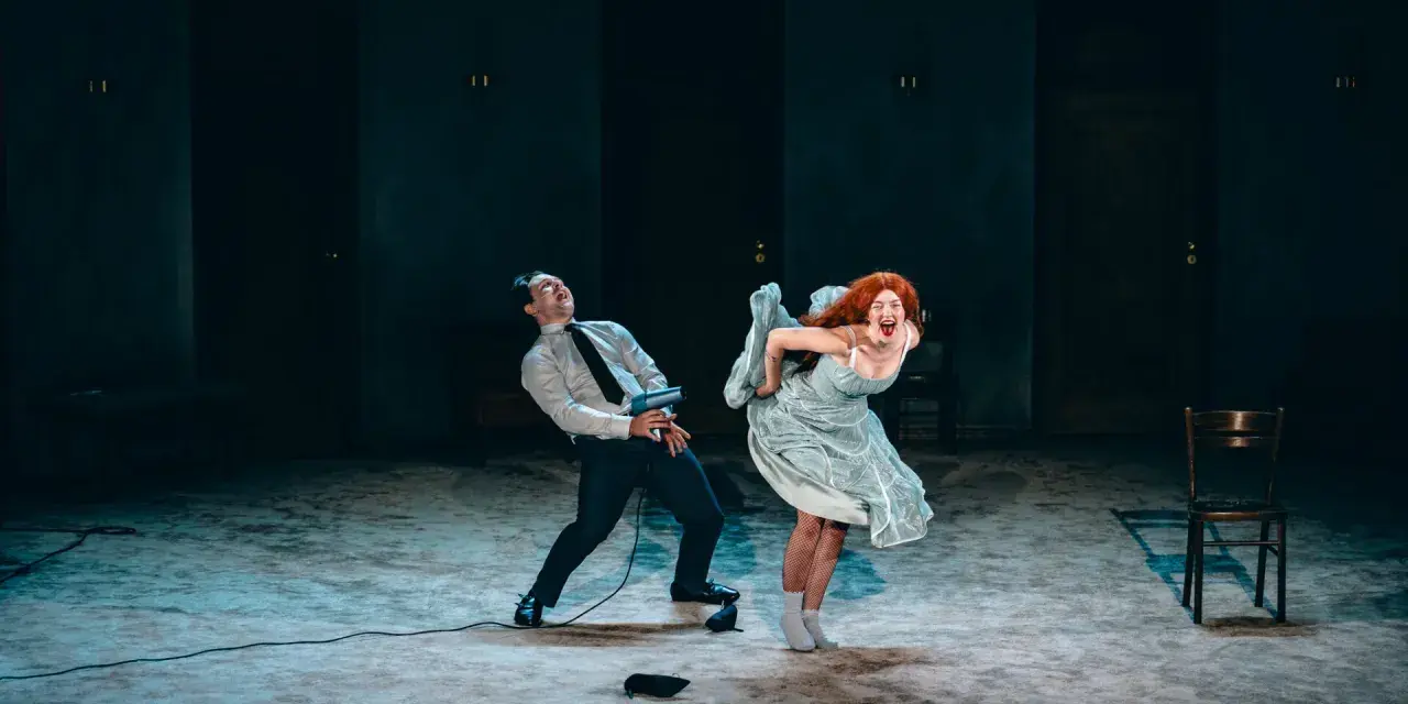 Dramatyczna scena z opery lub operetki: mężczyzna w garniturze i kobieta w zwiewnej sukni krzyczą, wyginając ciała w ekspresyjnych pozach.