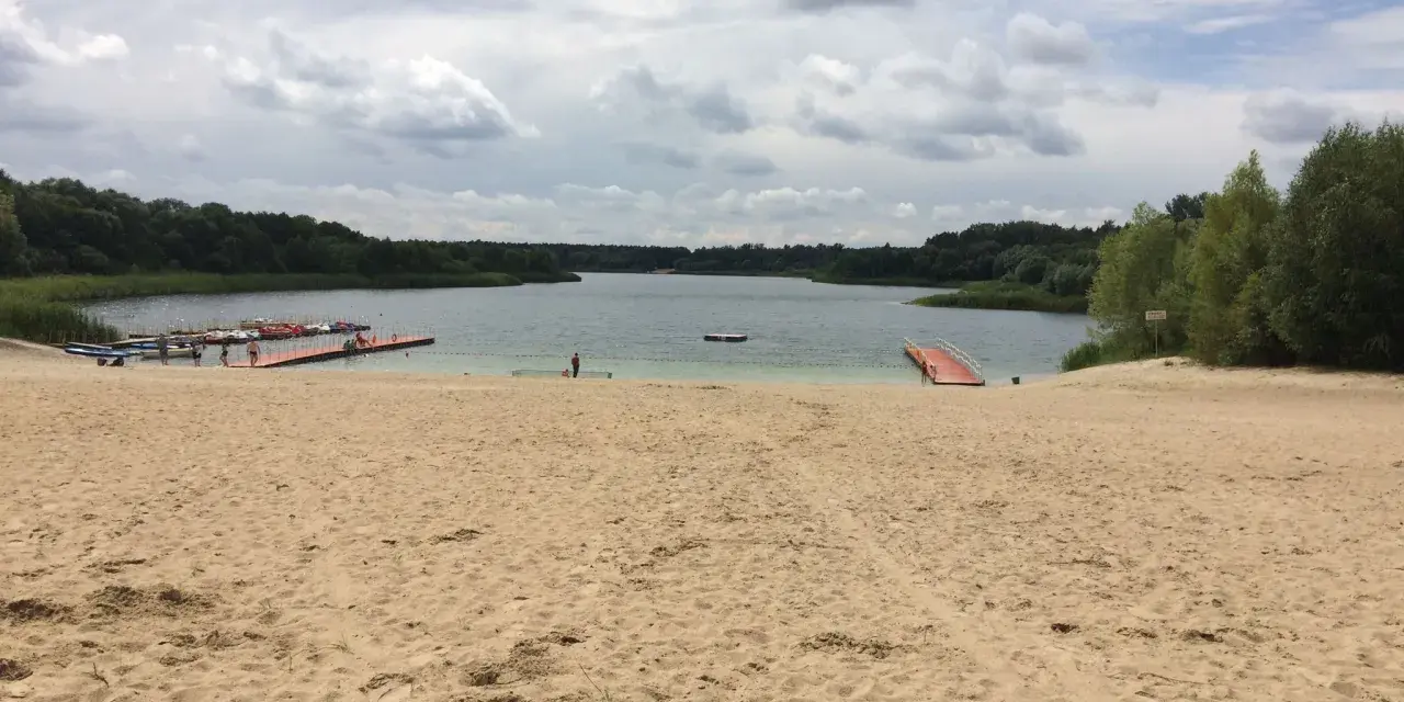 Piaskowa plaża nad jeziorem, z pomostami i łódkami. W oddali las, chmury na niebie. Idealne miejsce na wypoczynek nad wodą.
