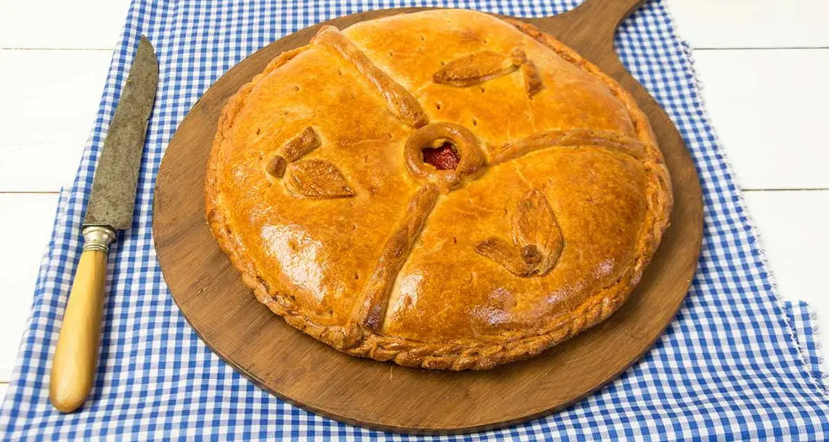 Empanada dorada y decorada sobre tabla de madera, con cuchillo y mantel a cuadros.