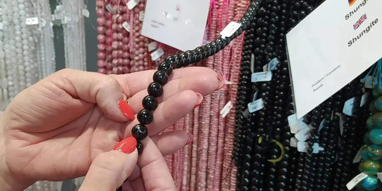 Manos con uñas rojas sostienen un collar de cuentas negras de shungita. En el fondo, se ven hilos de cuentas rosas y blancas.