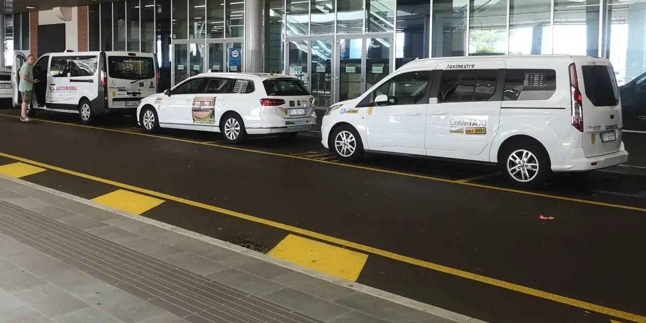 Taxi a Venezia, in attesa all'aeroporto. Diverse auto bianche, tra cui minivan e station wagon, pronte per il trasporto passeggeri.