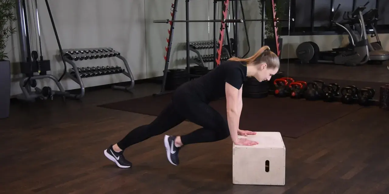 Kobieta wykonuje ćwiczenie mountain climbers na skrzyni. W tle siłownia z ciężarami.