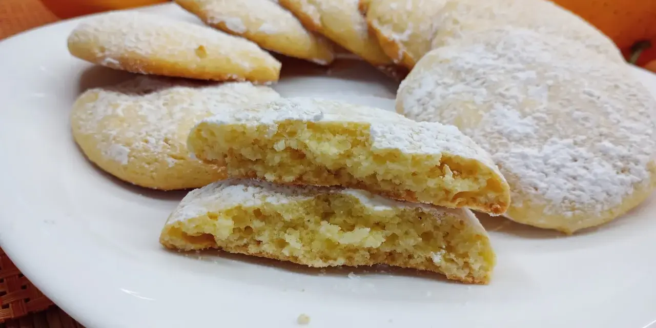 Biscotti morbidi all'arancia spolverati di zucchero a velo su un piatto bianco, con arance fresche sullo sfondo.