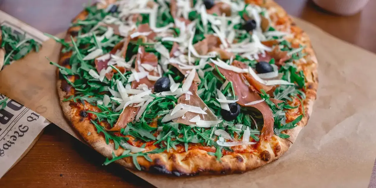 Pyszna pizza prosciutto z rukolą, oliwkami i parmezanem, podana na papierze do pieczenia.