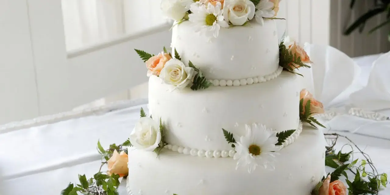 Elegancki tort weselny ozdobiony białymi i brzoskwiniowymi różami, margaretkami i zielonymi liśćmi. Piękne, jakie żywe kwiaty na tort!