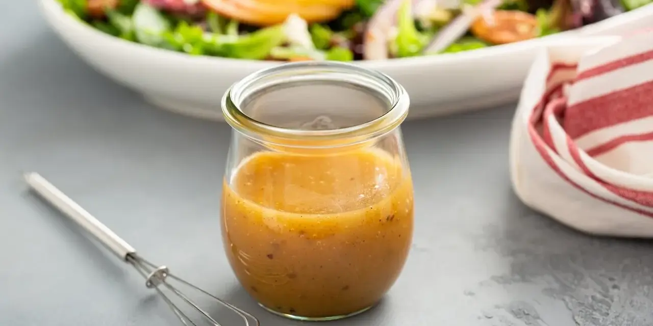 Ein Glas mit honig senf dressing steht neben einem kleinen Schneebesen. Im Hintergrund eine Schüssel mit Salat.