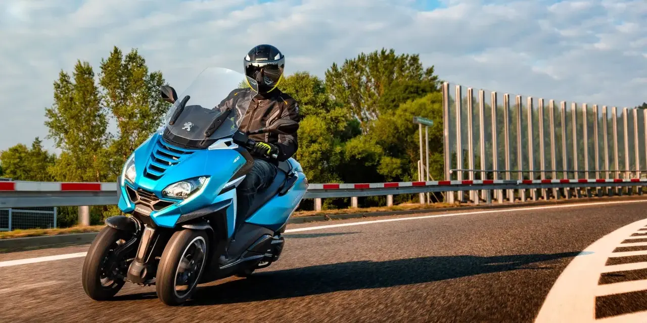 Niebieski Peugeot Metropolis 400 z kierowcą w kasku pędzi po zakręcie drogi, otoczony zielenią i błękitnym niebem.