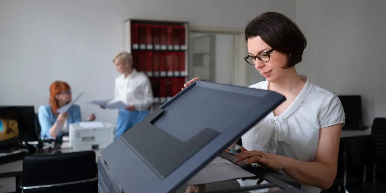 Frau mit Brille bedient einen Drucker, vielleicht um einen Epson Drucker zu installieren. Im Hintergrund arbeiten Kolleginnen.