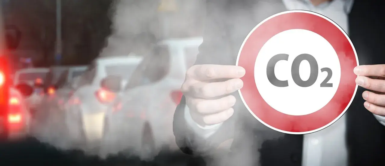 Dłoń trzyma znak zakazu z napisem CO2 na tle dymiących samochodów w korku.