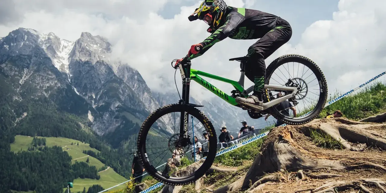 Troy Brosnan in mid-air on a green downhill bike, with a dramatic mountain backdrop.